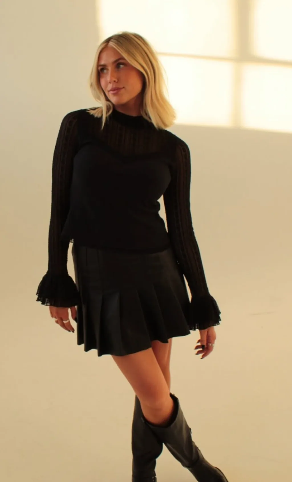 Graci Mortensen in a black lace top and pleated skirt, standing in a sunlit studio